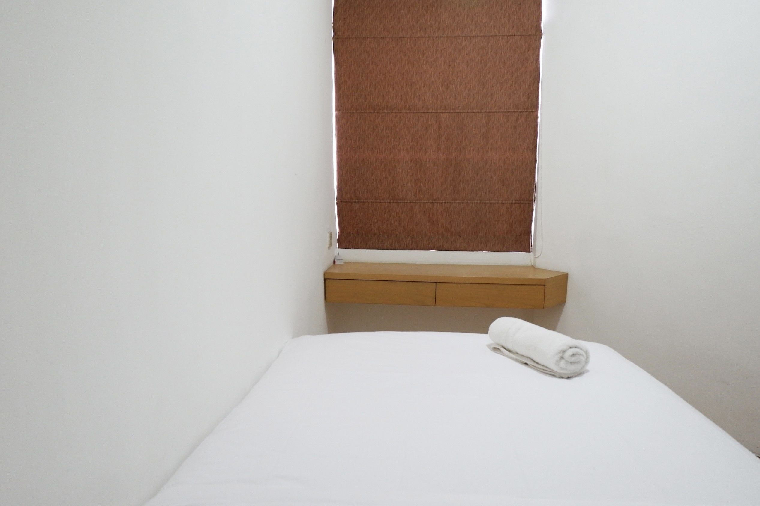 A single bed with a white mattress and a rolled white towel on it. There is a wooden bedside table with a drawer and a small shelf. A window with a brown blind is visible above the bed.