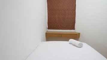 A single bed with a white mattress and a rolled white towel on it. There is a wooden bedside table with a drawer and a small shelf. A window with a brown blind is visible above the bed.