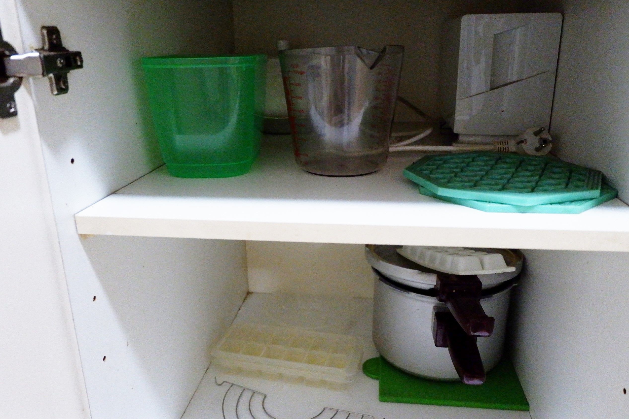 A kitchen shelf with a green plastic container, a measuring cup, a green cutting board, and a white appliance.