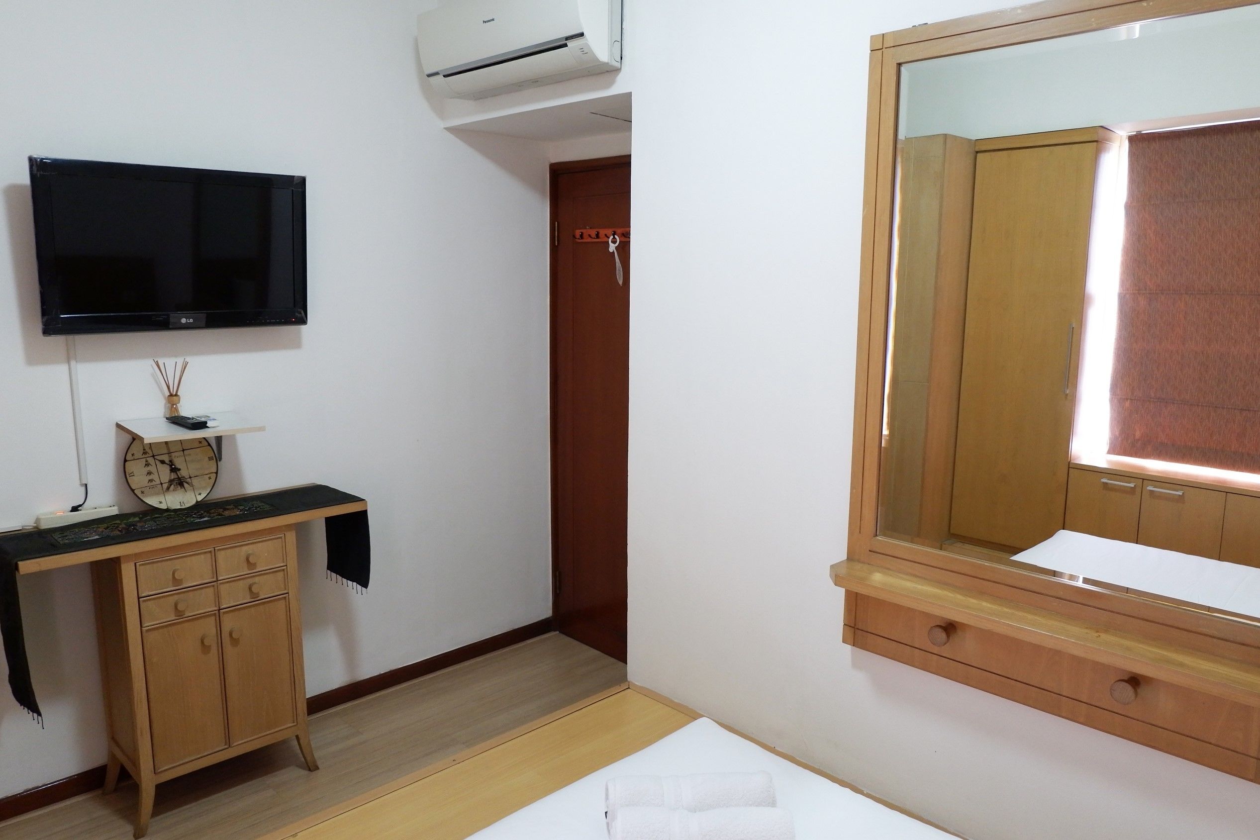 Una habitación con un armario de madera, un televisor de pantalla plana montado en la pared, un escritorio de madera con un reloj y un espejo grande sobre el armario.
