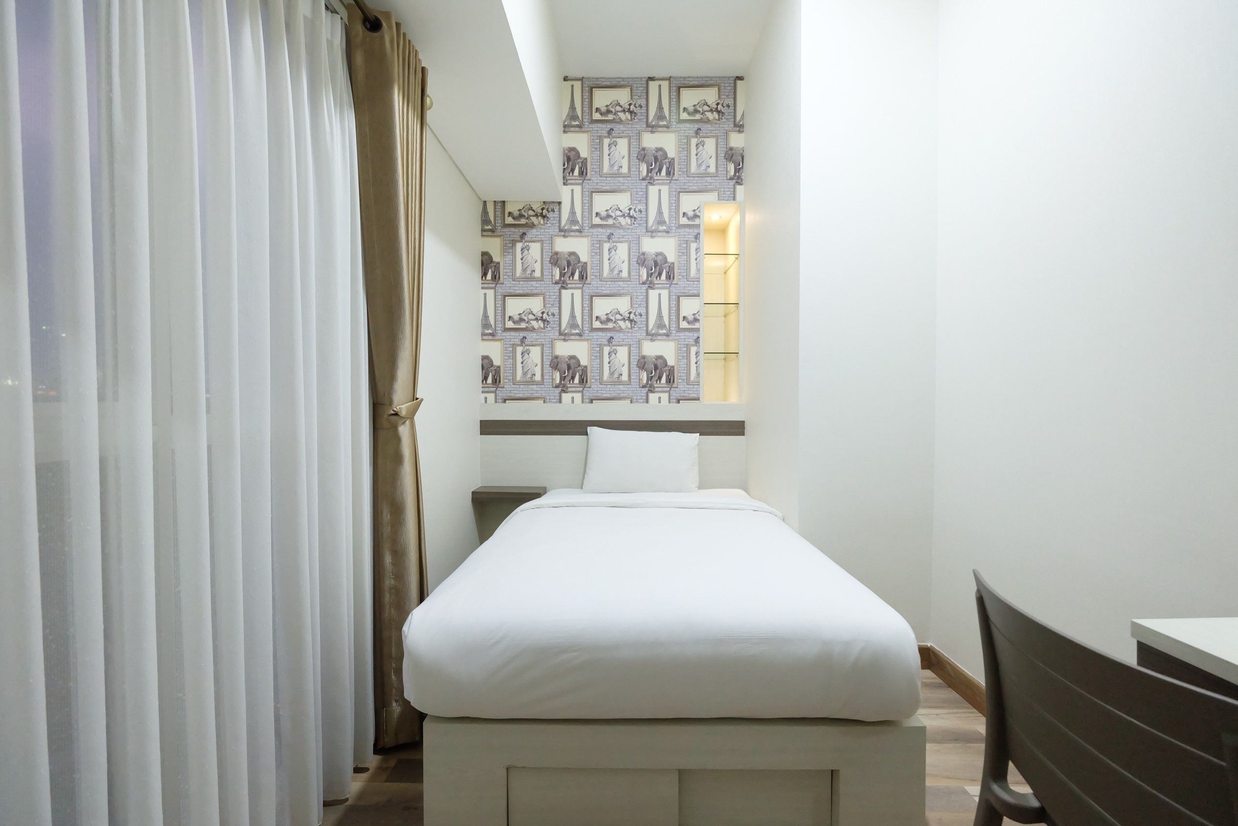 A small, neatly arranged bedroom with a bed, a nightstand, a chair, and a wall with framed pictures.