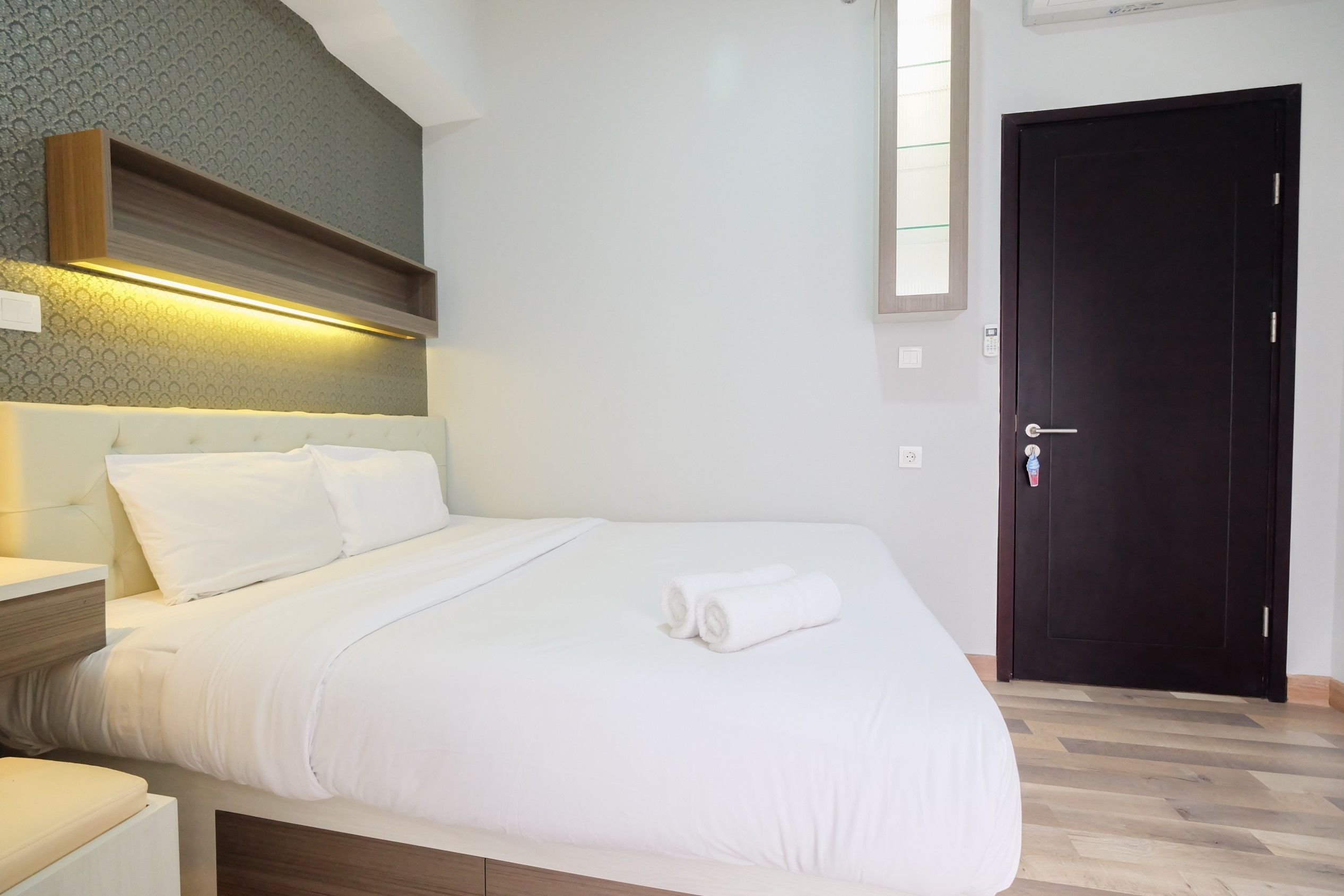 A neatly made bed with white bedding and a pair of rolled towels, a bedside table with a lamp, and a dark door in a hotel room.