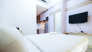 A hotel room with a bed, a TV mounted on the wall, and a kitchenette in the background.