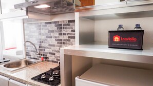 A kitchen with a gas stove, a sink, and a backsplash of gray tiles. There is a shelf with a box labeled "Travelio" and two water bottles on it. - Brand New Studio Room Cinere Bellevue Suites Apartment (Depok)