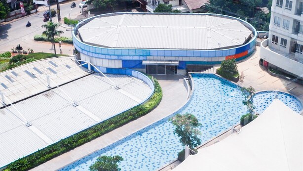A large building with a circular blue and white facade, adjacent to a swimming pool with a blue tiled pattern. - Brand New Studio Room Cinere Bellevue Suites Apartment (Depok)