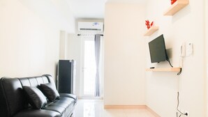 A modern living room with a black leather sofa, a flat-screen TV mounted on a wooden shelf, and a large window with curtains. - Simply Minimalist 2BR The Springlake Apartment (Bekasi)