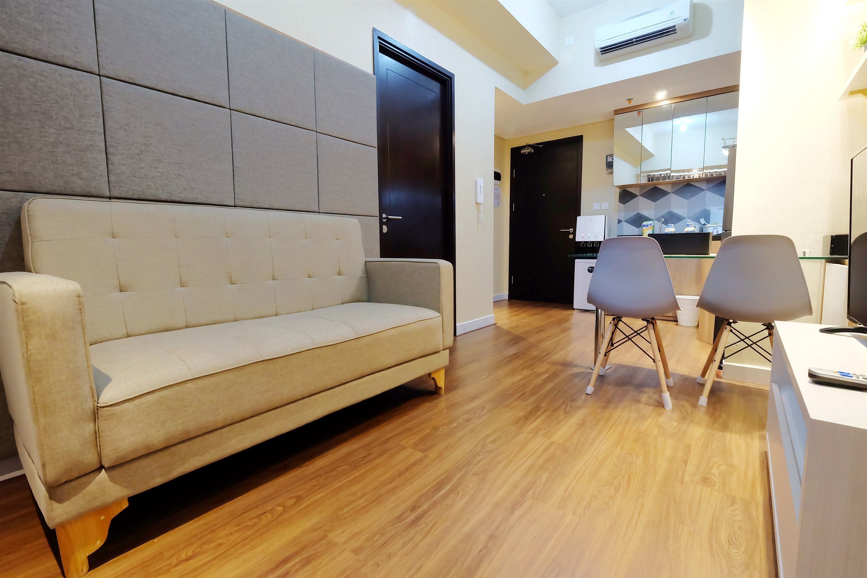 A modern living room with a beige sofa, wooden flooring, a black door, and a kitchen area visible through an open door.