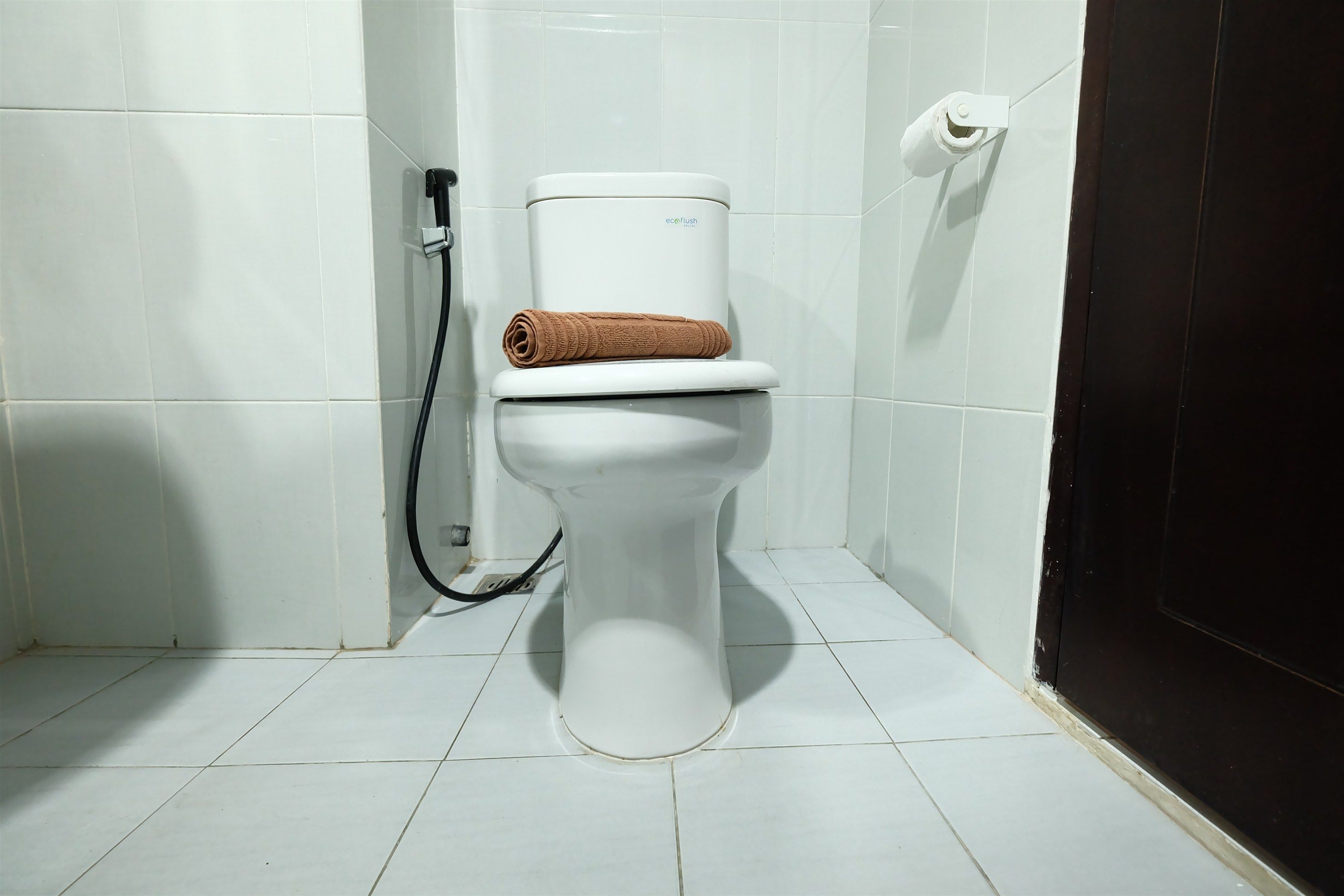 A white toilet with a brown towel on top, a toilet paper holder, and a black hose.