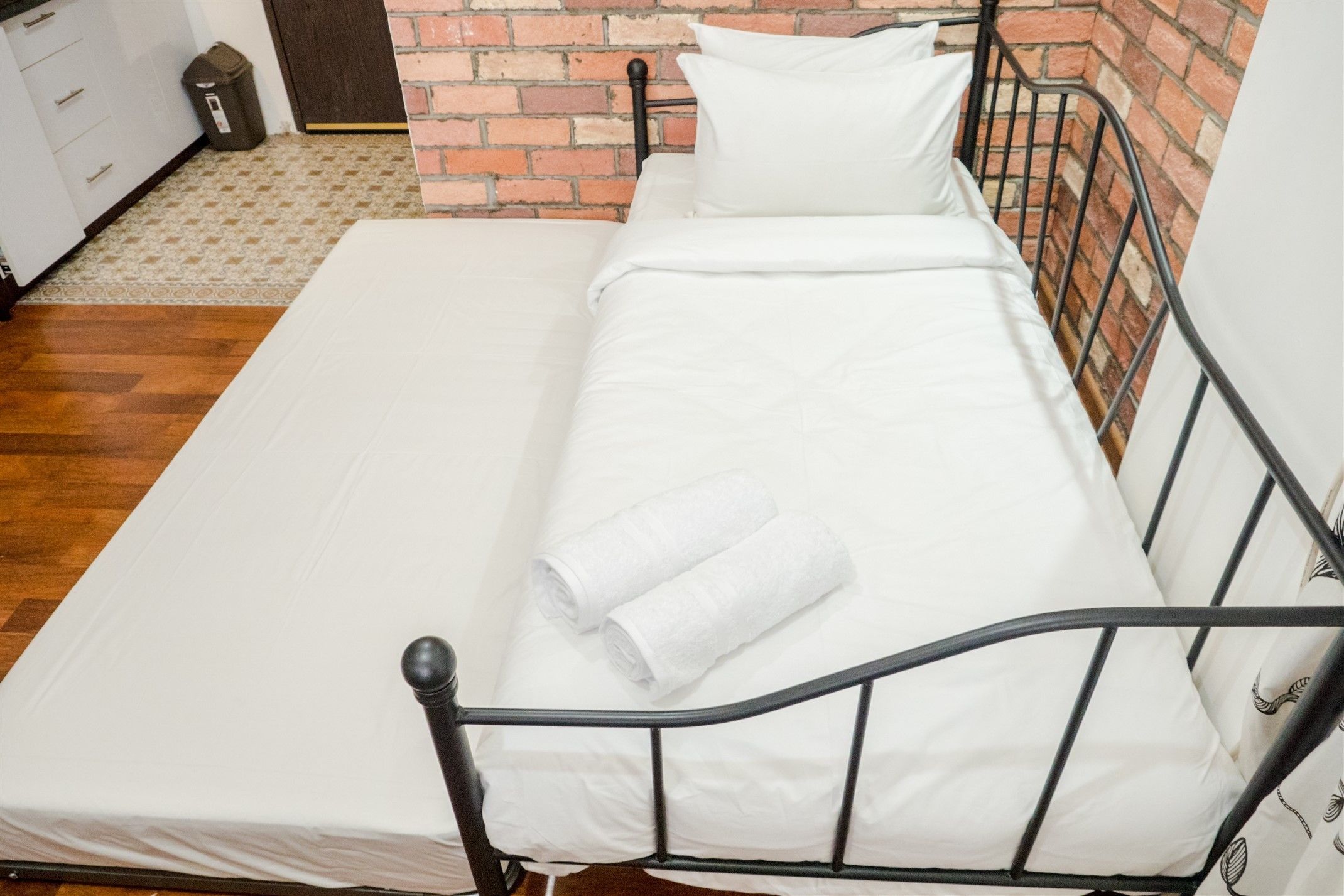 Uma cama de solteiro com estrutura de metal, roupa de cama branca e uma toalha enrolada.