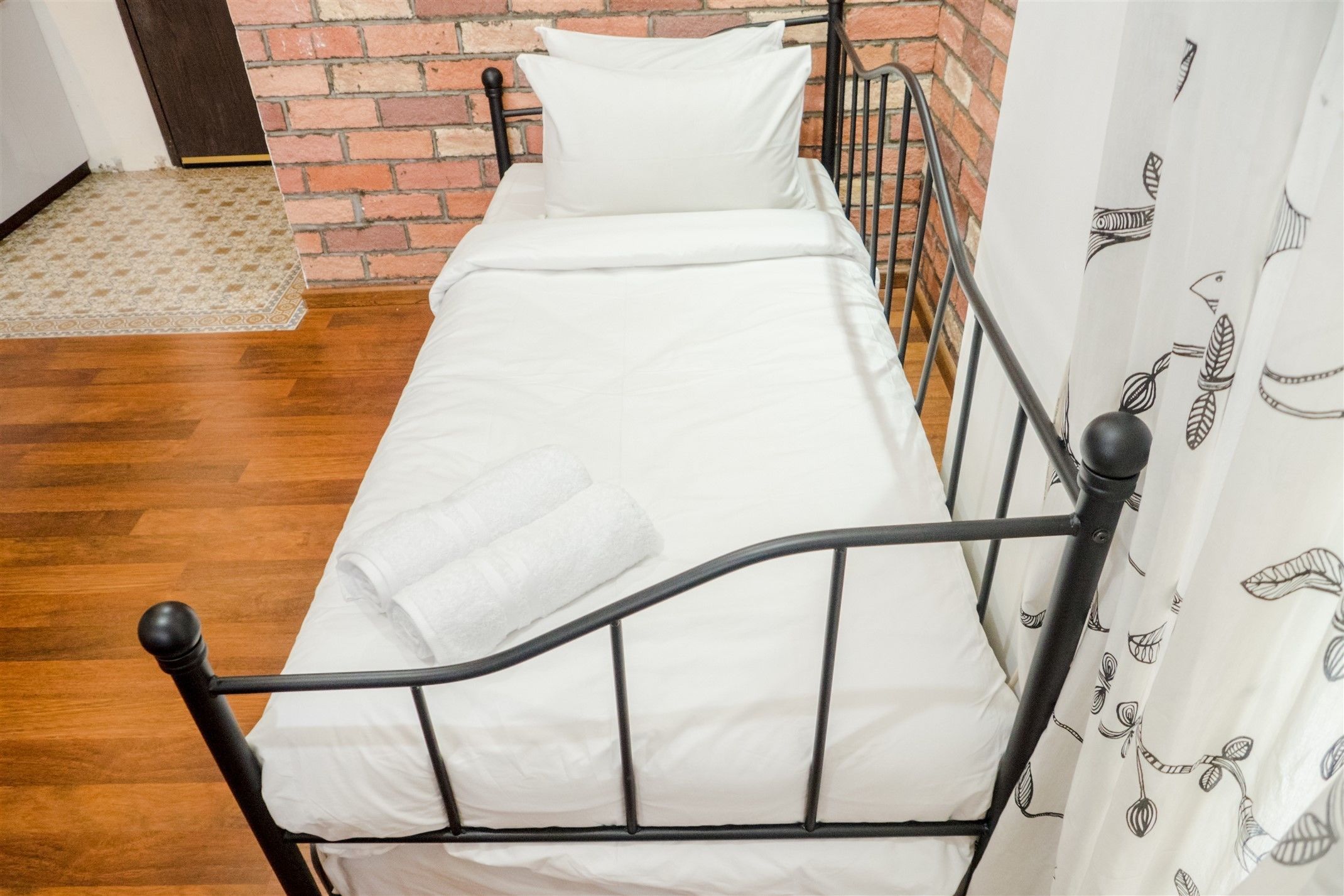 Ein Einzelbett mit weißer Bettwäsche, einem schwarzen Metallgestell und einem weißen Vorhang mit Blätermotiven.