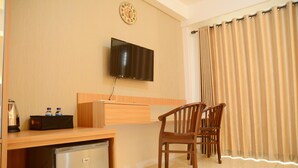 A compact living space with a flat-screen TV mounted on the wall, a wooden desk with a small refrigerator, two wooden chairs, and a large window with curtains. - Simply Studio Room @ Annora Living Apartement Tangerang (Tangerang)