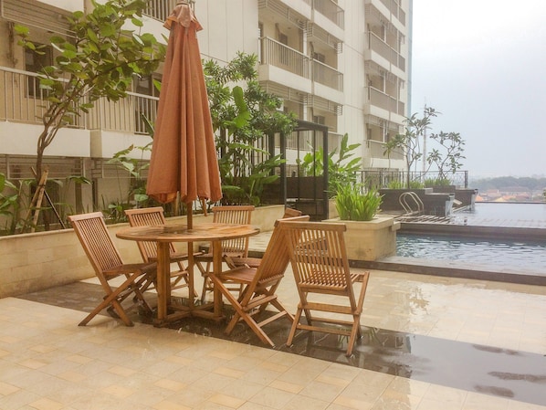 A patio with a round wooden table and chairs, a brown umbrella, and a pool in the background.