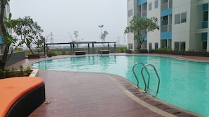 A swimming pool with a wooden deck, lounge chairs, and a view of a building with balconies. - Homey Studio Room The Nest Apartment (Tangerang)