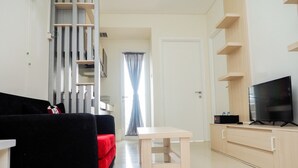 A modern living room with a black sofa, a small wooden table, a TV on a wooden cabinet, and a staircase leading to another level. - Comfortable 2BR Parahyangan Residence Apartment (Bandung)