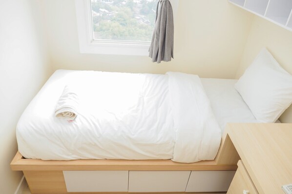 A single bed with a white mattress and pillows, a wooden headboard, and a window with a view of greenery.