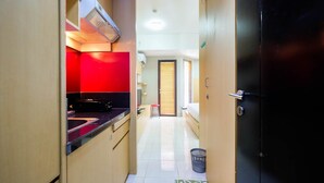 A narrow hallway with a kitchen area on the left, featuring a red backsplash and a black countertop. A black door is on the right, and a small trash bin is on the floor. - Central Jakarta Studio Apartment At Tamansari Sudirman (Jakarta)