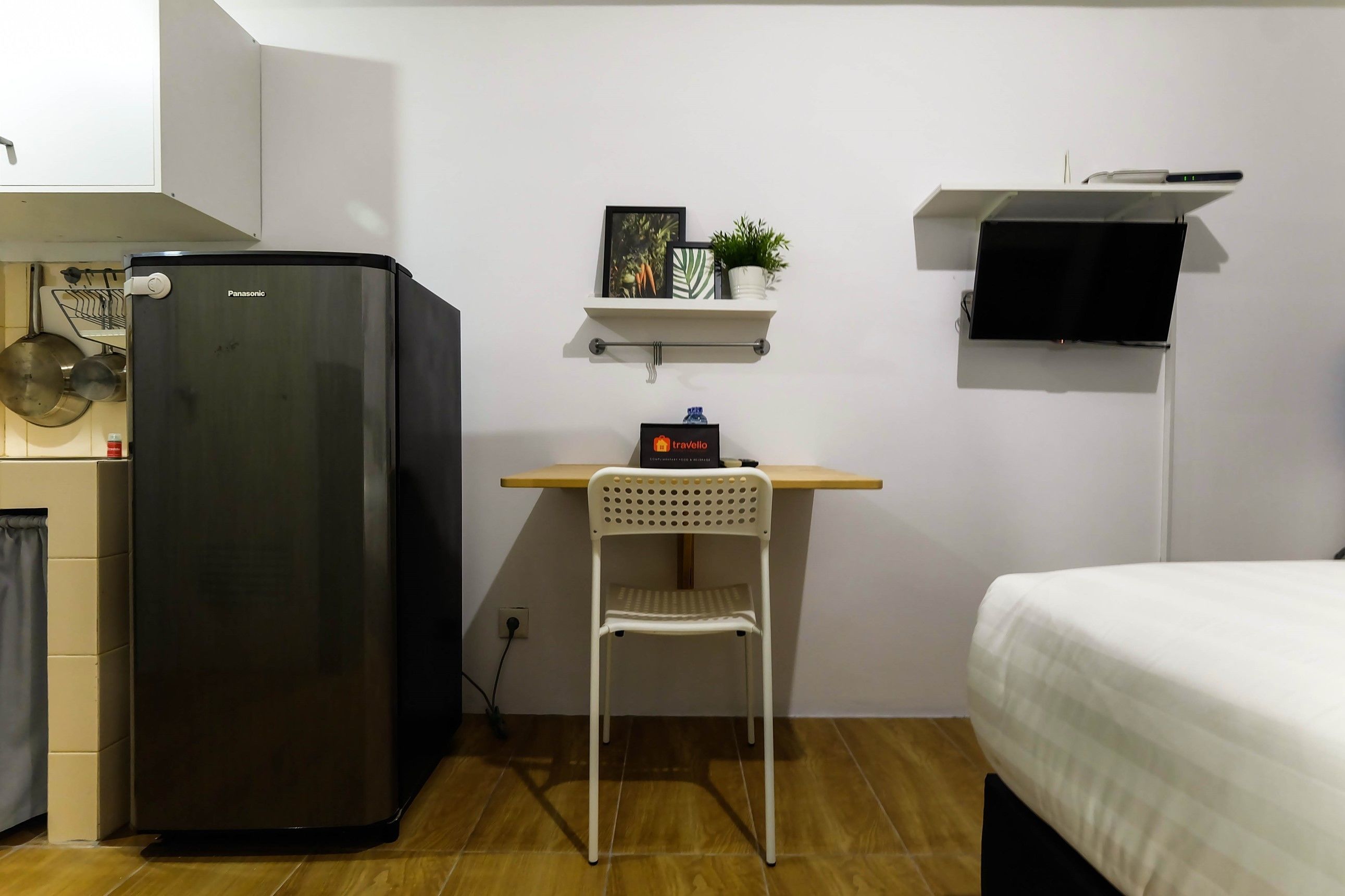 A compact room with a black refrigerator, a white chair, a bed, a shelf with a plant, and a mounted TV.