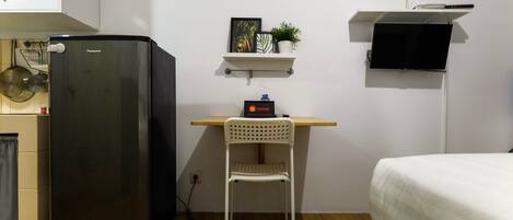 A compact room with a black refrigerator, a white chair, a bed, a shelf with a plant, and a mounted TV.
