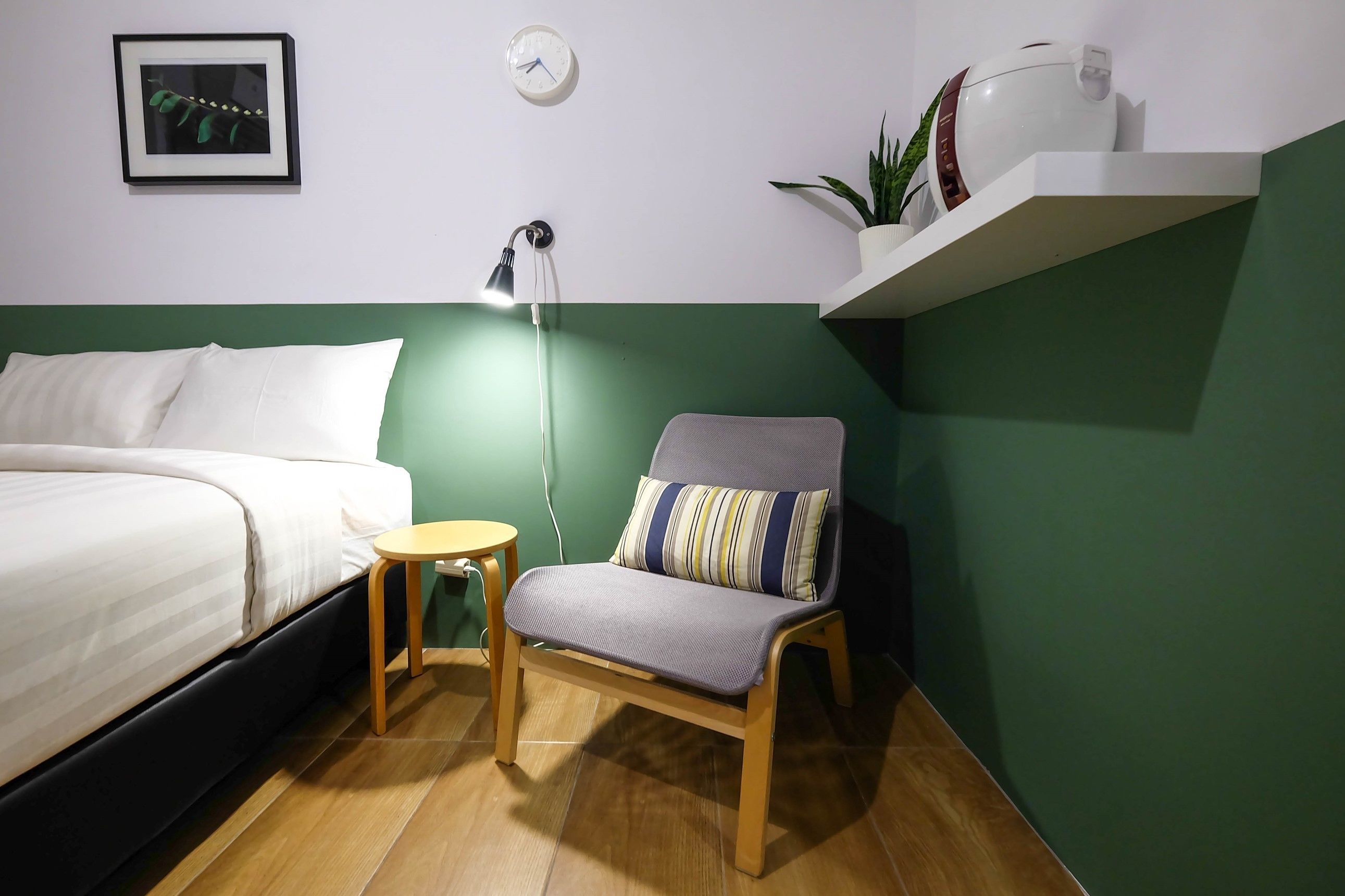 A neatly arranged bedroom with a bed, a chair, a small table, a wall clock, and a shelf with a plant.