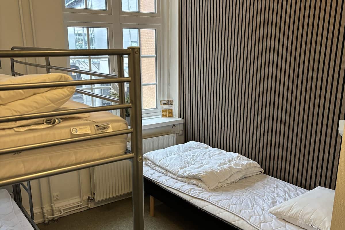 A dormitory room with bunk beds, a single bed, a radiator, and a window with a view of buildings.