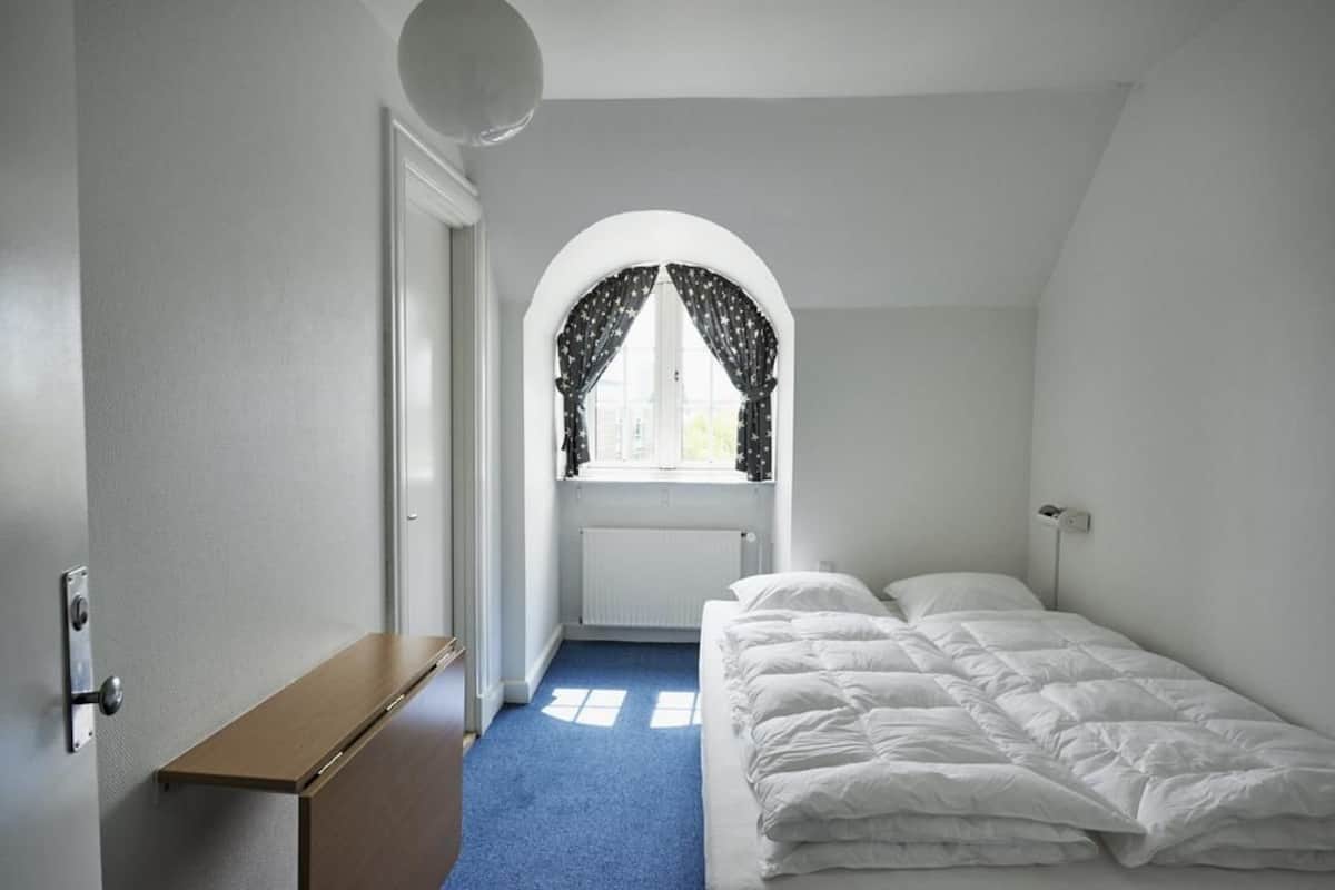 A bedroom with a single bed, a window with star-patterned curtains, a radiator, and a wooden bench.
