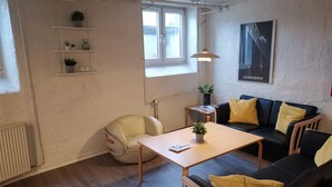 A modern living room with a black leather sofa, a wooden coffee table, and a white armchair. There are shelves with plants and a radiator.