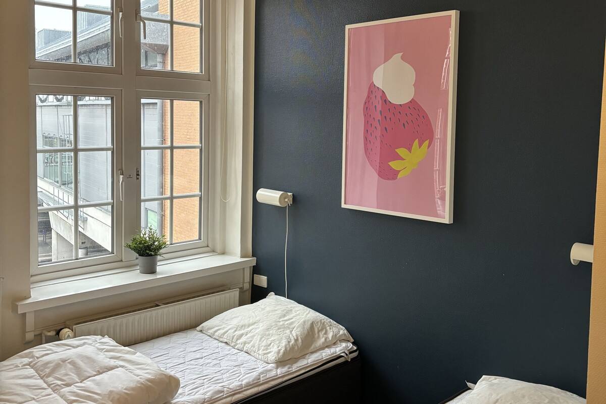 A bedroom with two beds, a window with a view of buildings, a radiator, and a wall-mounted light fixture.