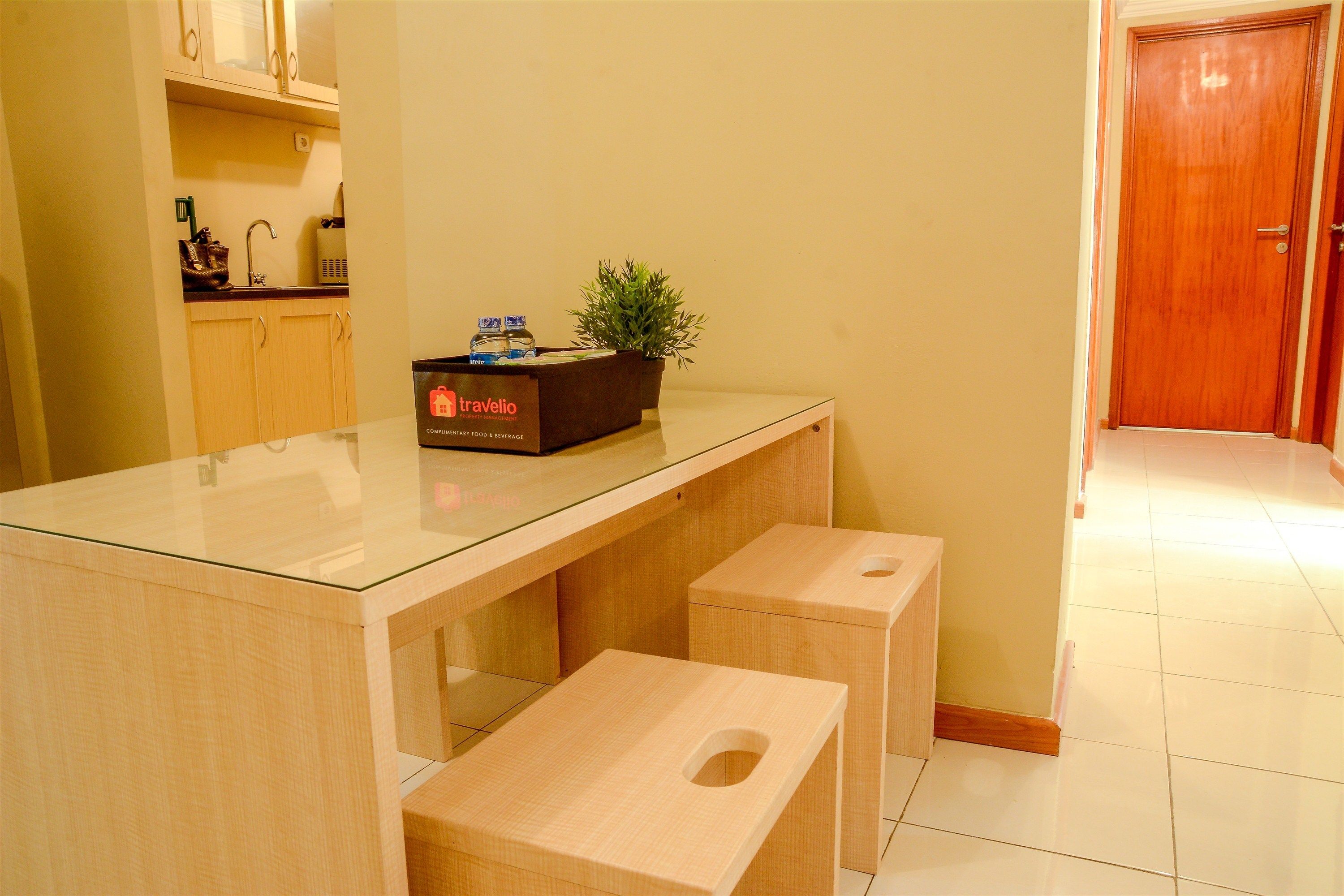 A clean, modern kitchen with a glass-top table, wooden stools, a box labeled 'travello', and a potted plant.
