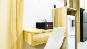 A compact room with a wooden desk, a white chair, a black box labeled 'Travelio', and a white refrigerator. - Modern Style The Oasis Studio Apartment with Comfortable Sofa (Cikarang)