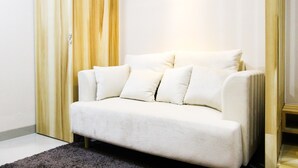 A white sofa with multiple cushions in a room with wooden paneling and a small wooden table. - Modern Style The Oasis Studio Apartment with Comfortable Sofa (Cikarang)