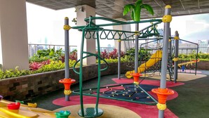 Un espace de jeux extérieur doté de diverses structures ludiques, notamment un toboggan et des jeux d’escalade, entouré de tapis de jeu colorés et de plantes en pot.