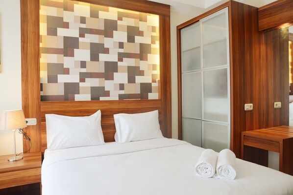 A neatly made bed with white linens and two rolled towels, a wooden headboard, a wall with a geometric pattern, and a wooden nightstand with a lamp.