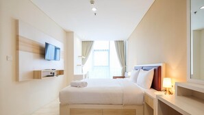 A modern hotel room with a large bed, a flat-screen TV mounted on the wall, a bedside table with a lamp, and a window with curtains. - 2BR Cozy and Modern L'Avenue Apartment (Jakarta)
