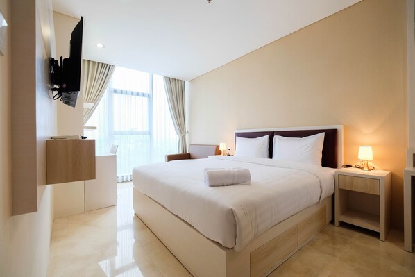 A modern hotel room with a large bed, a TV mounted on the wall, a bedside table with a lamp, and a view of the outdoors through large windows. - 2BR Cozy and Modern L'Avenue Apartment (Jakarta)