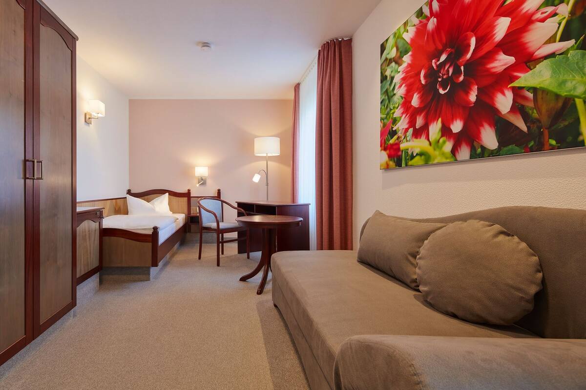 A hotel room with a bed, a desk, a chair, a sofa, and a large floral painting on the wall.