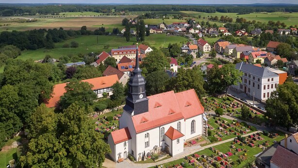 An aerial view of a village with a prominent church featuring a tall steeple, surrounded by residential buildings and a well-maintained cemetery. - Hotel garni Sonnenhof (Moritzburg)