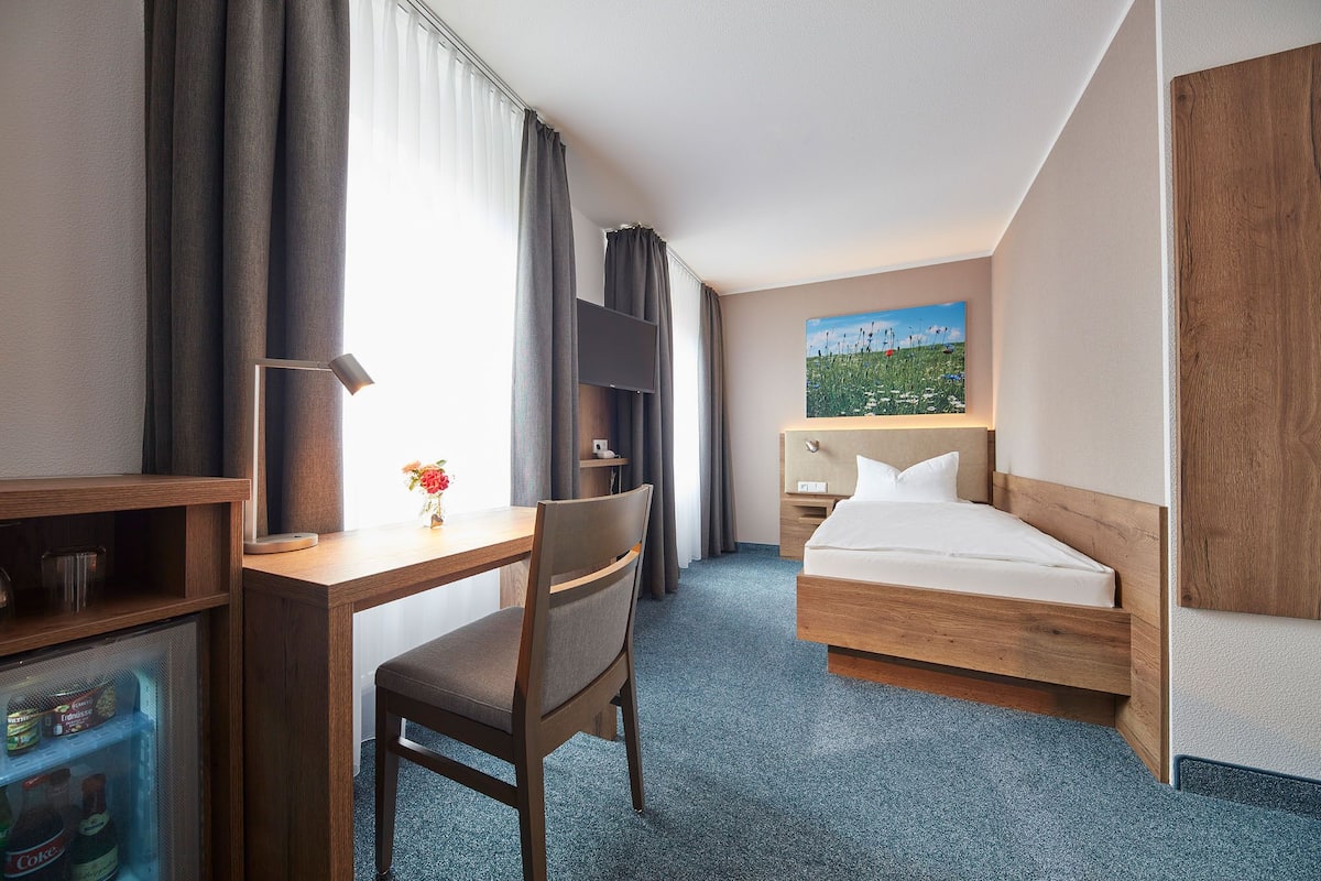 A hotel room with a bed, a desk, a chair, a TV, and a painting on the wall.