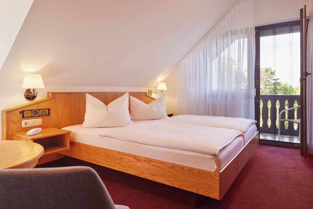 A hotel room with a wooden bed, two bedside lamps, a bedside table with a phone, and a view of the outdoors through a sliding glass door.