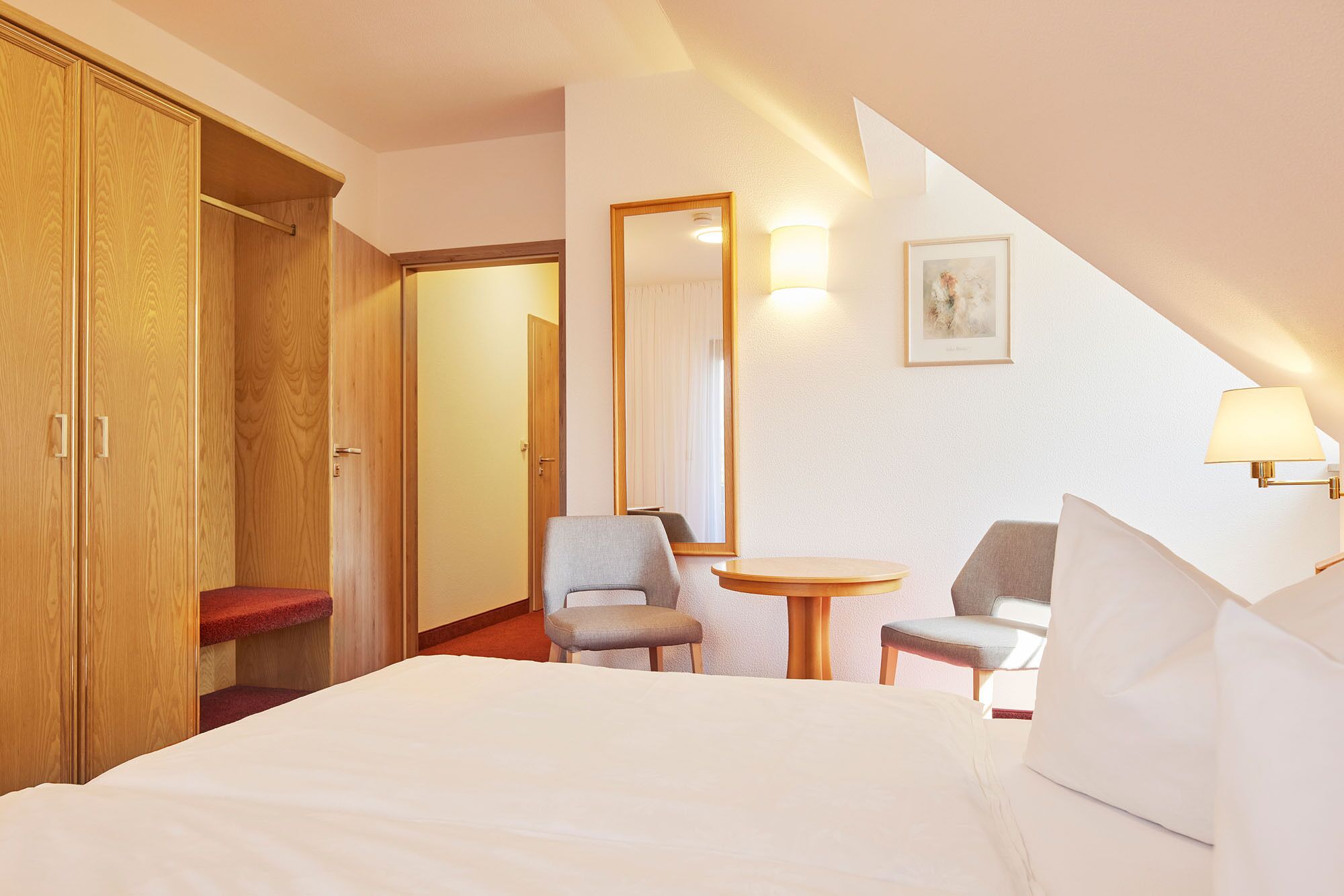 A hotel room with a bed, two chairs, a small table, a mirror, and a wooden wardrobe.
