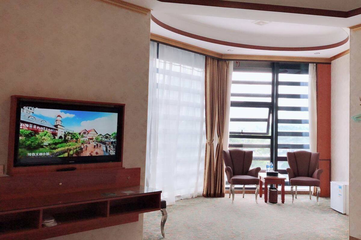 A hotel room with a TV displaying a scenic view, two armchairs, a small table, and a large window with curtains.