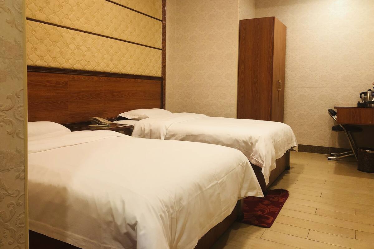 A hotel room with two beds, a wooden wardrobe, a desk with a chair, and a patterned wall.