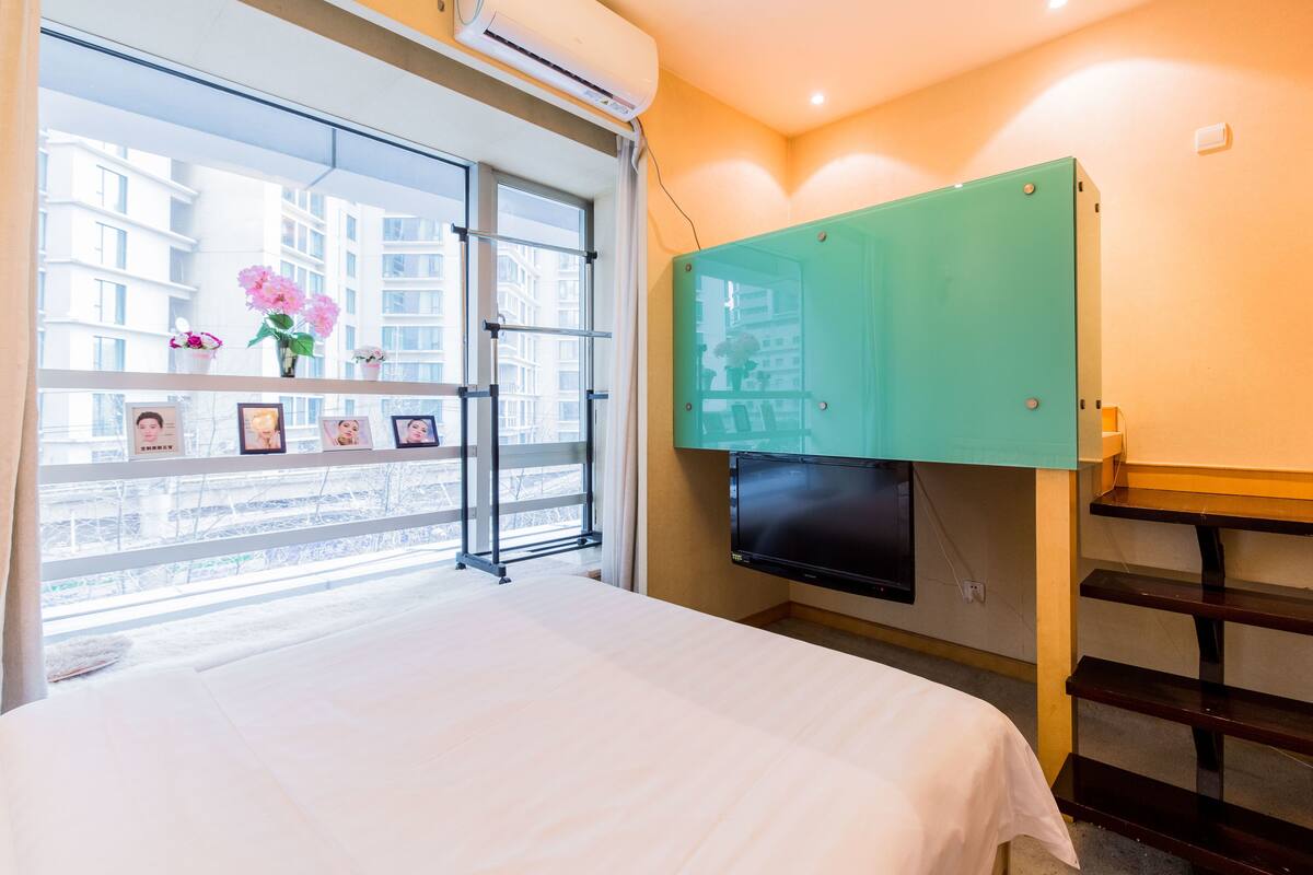 Habitación de hotel con cama, televisión montada en la pared, ventana con vistas a edificios y estante con flores y fotografías.