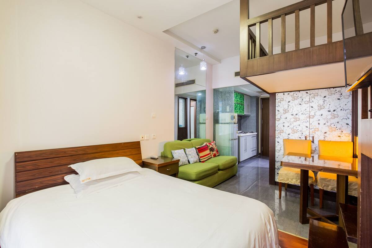Habitación de hotel con cama, sofá verde, mesita con lámpara y escalera que lleva a un nivel superior.