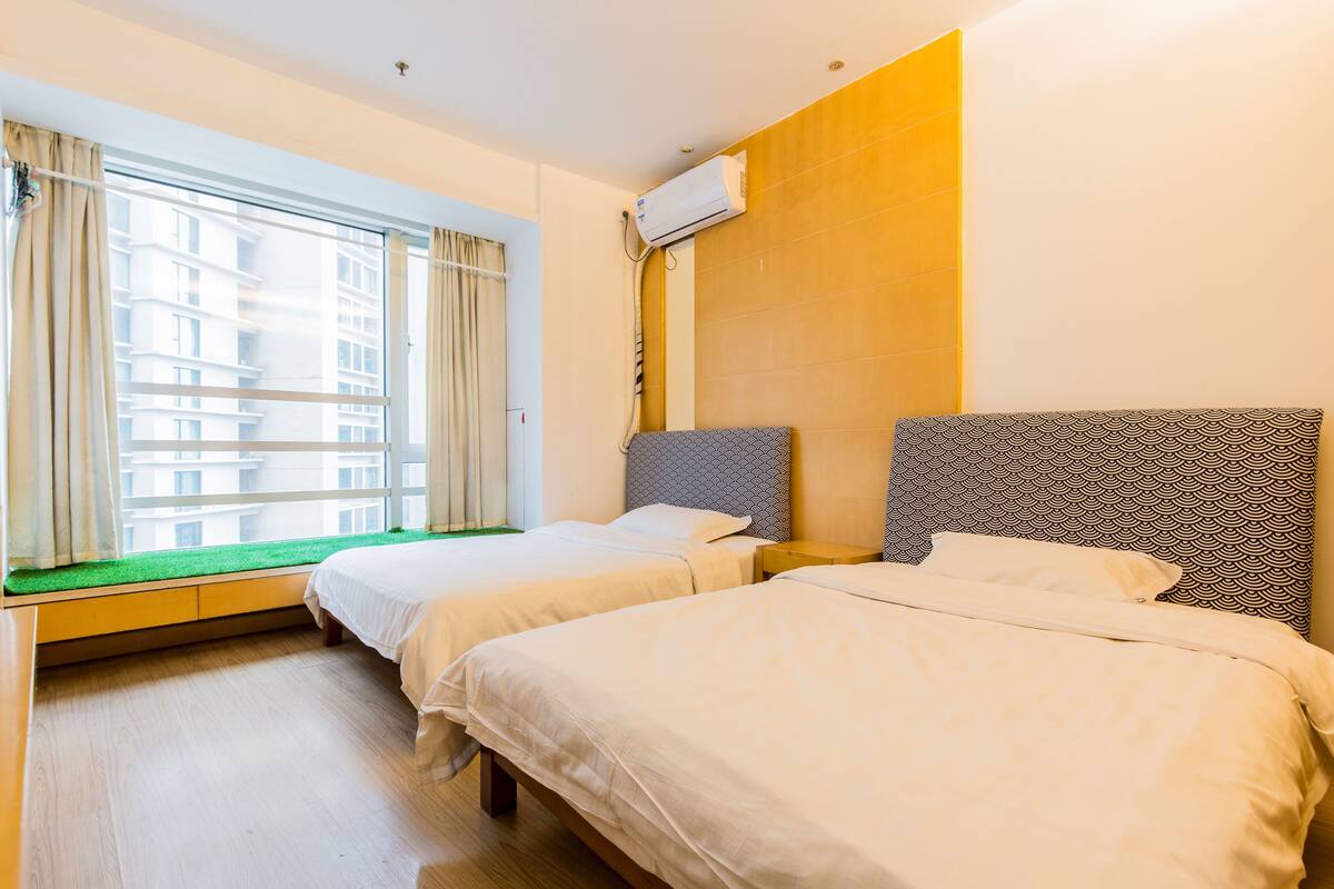 Habitación de hotel con dos camas, una ventana con cortinas, un banco empotrado y un equipo de aire acondicionado.