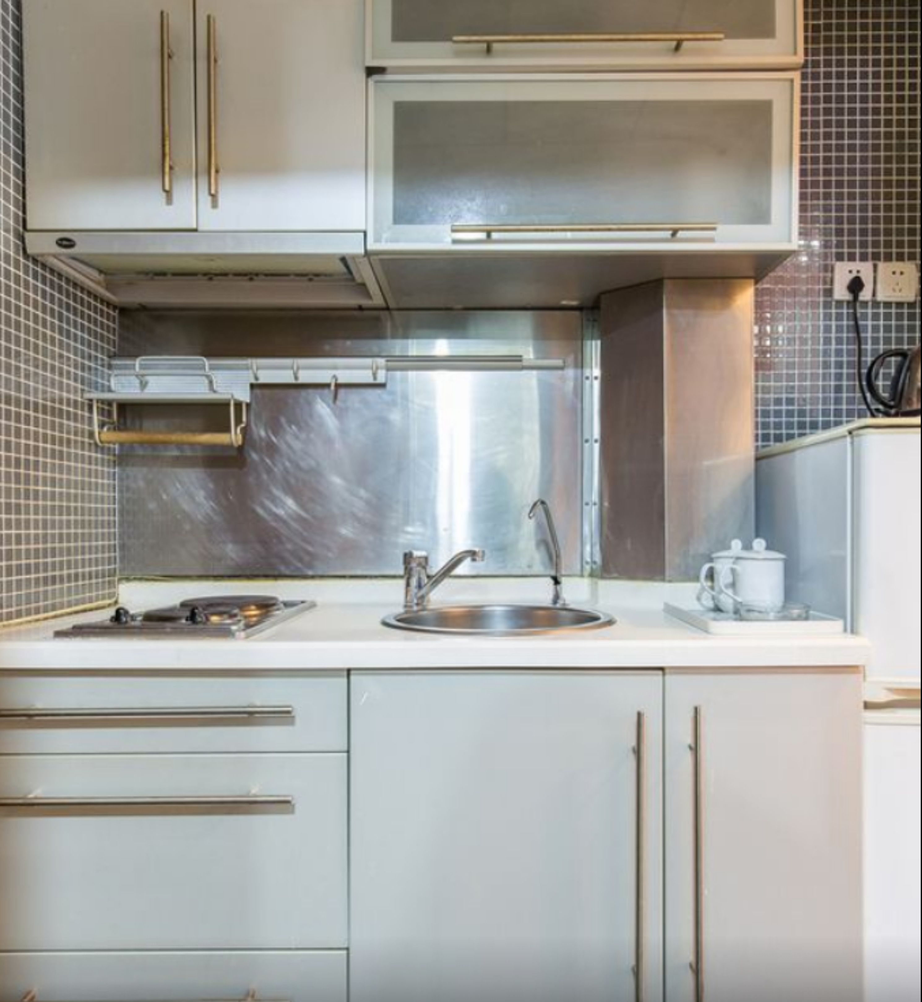 Una cocina con un lavamanos de acero inoxidable, un microondas y una estufa. Hay armarios arriba y abajo del mostrador, y un salpicadero de azulejos.