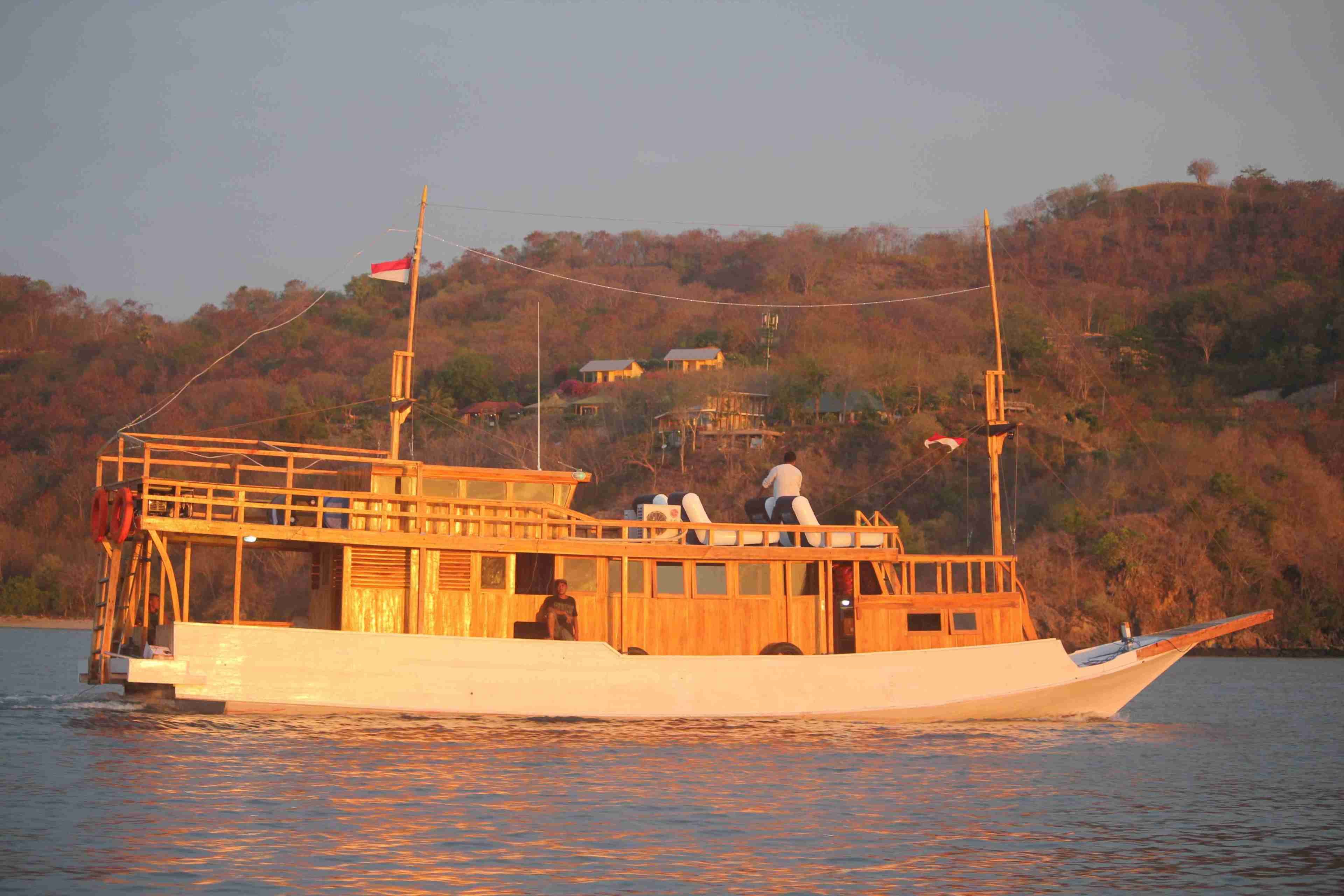 A wooden boat with a cabin and deck seating, sailing on calm water with a forested hillside in the background.