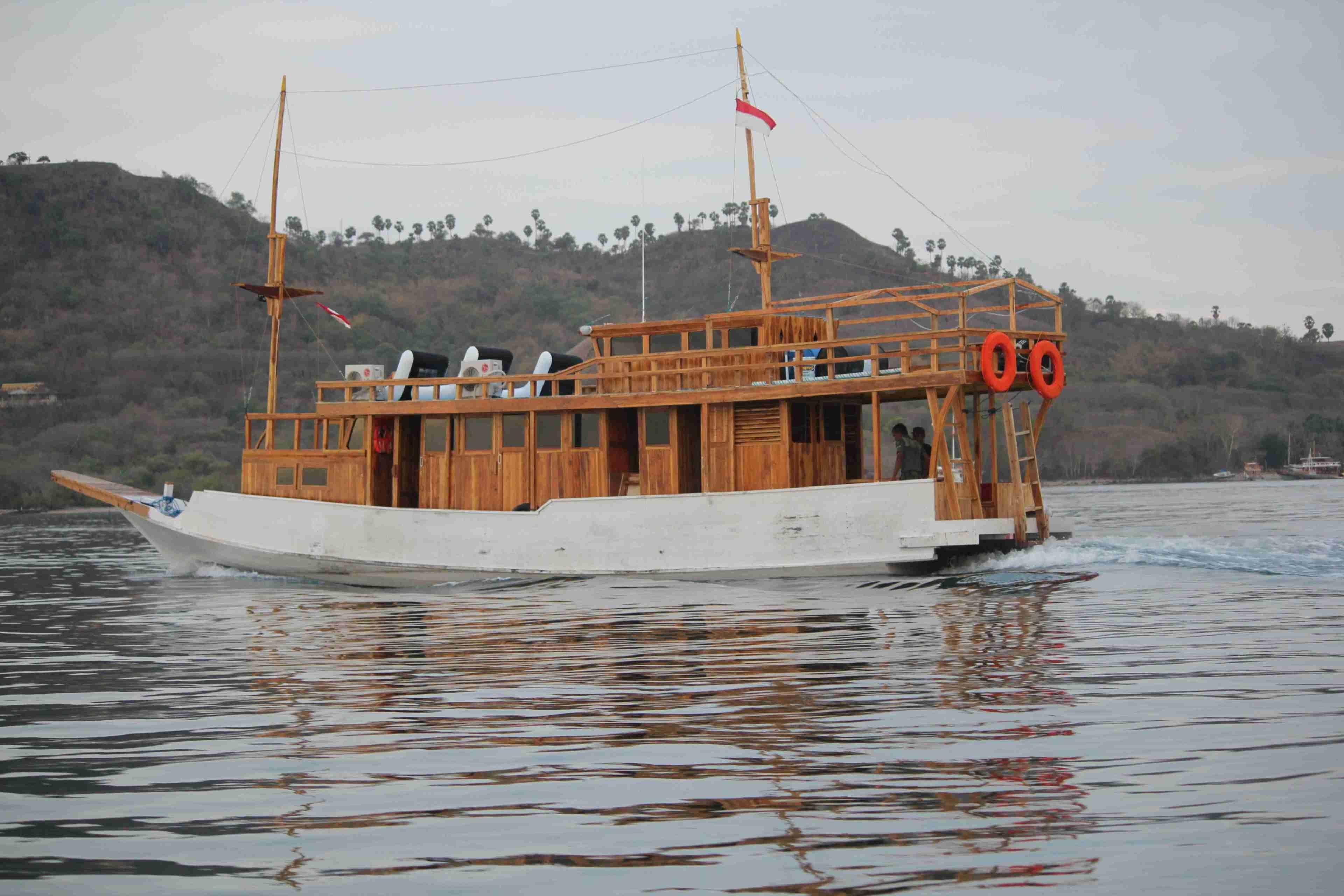 A wooden boat with a cabin and two masts, moving on water with a forested hillside in the background.