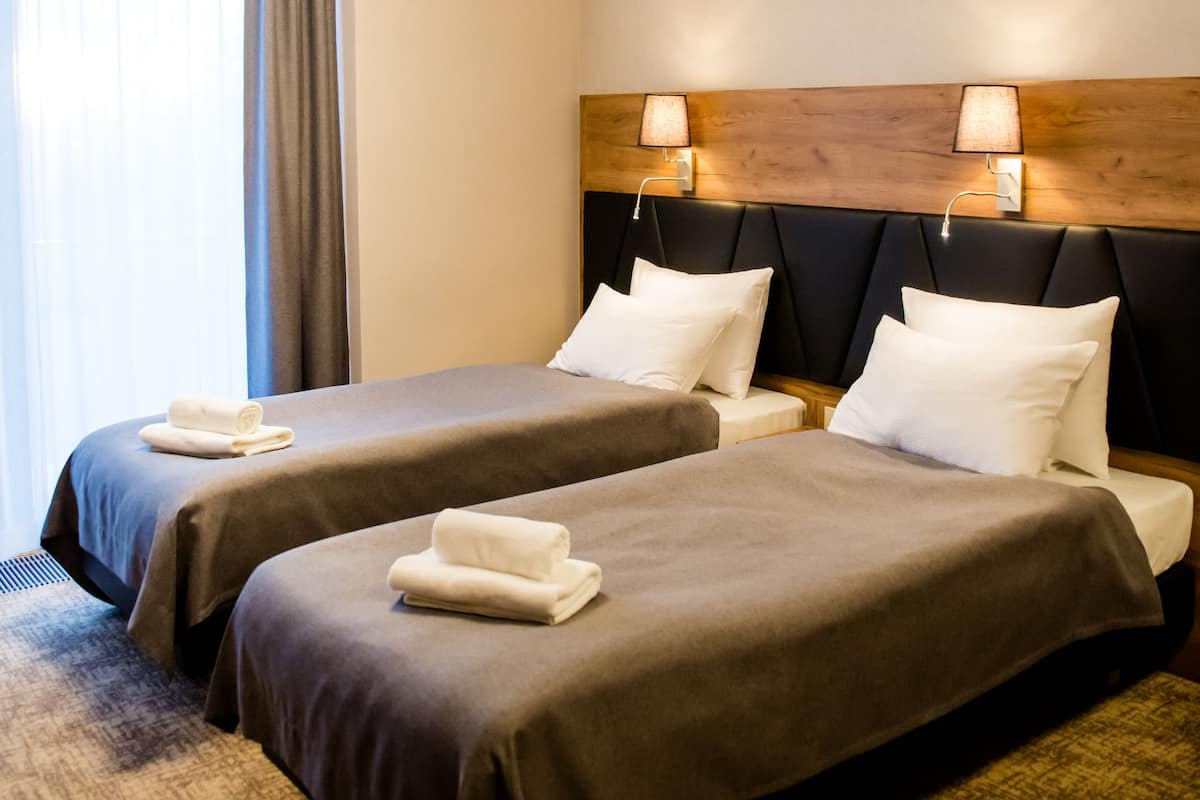 A hotel room with two beds, a headboard, bedside lamps, a window with curtains, and an air conditioning unit.