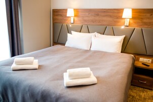 Cama bem arrumada com duas toalhas dobradas, cabeceira de madeira com duas luminárias fixadas na parede e mesa de cabeceira com um controle remoto.