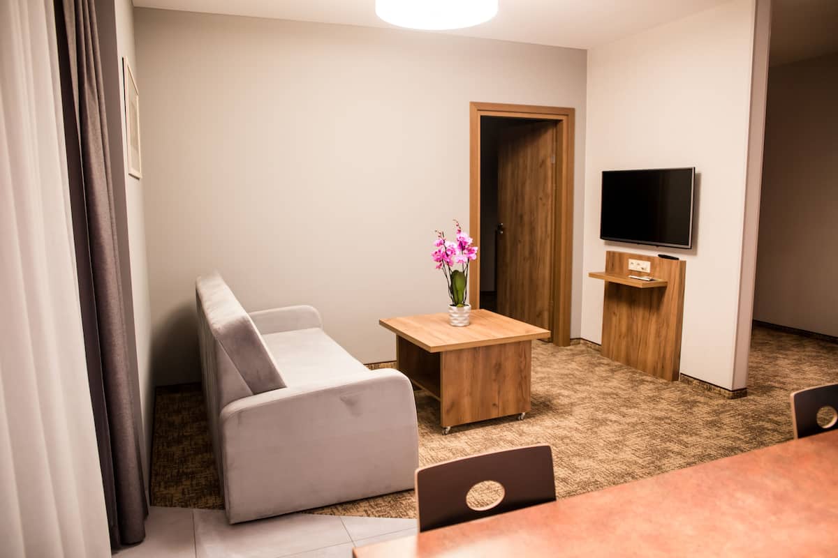 A modern living room with a grey sofa, a wooden coffee table with a vase of flowers, a flat-screen TV on a wooden stand, and a laptop on a table.